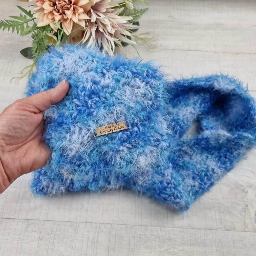 Blue textured fabric held by a hand with a 'Country Crafts' label, on a light wooden surface with flowers in the background.