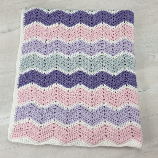 Crocheted blanket with a chevron pattern in purple, pink, and white on a wooden surface.
