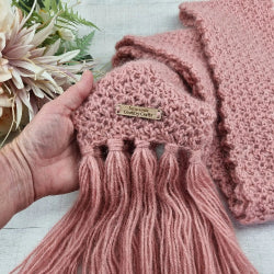 Pink knitted item with tassels held by a hand, next to a pink blanket and flowers on a light surface.