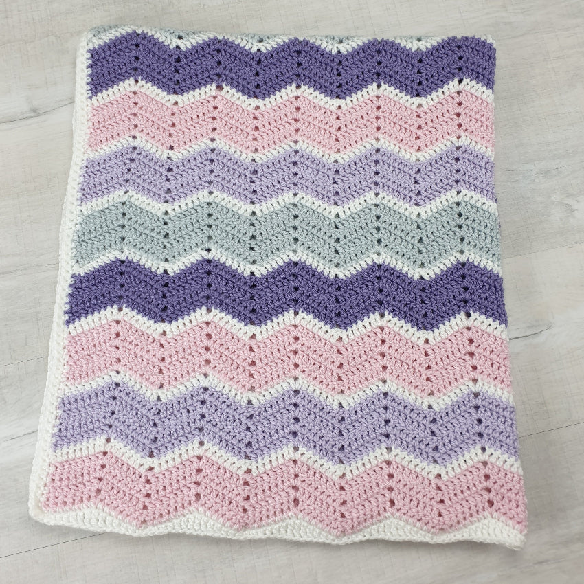 Crocheted blanket with a chevron pattern in purple, pink, and white on a wooden surface.